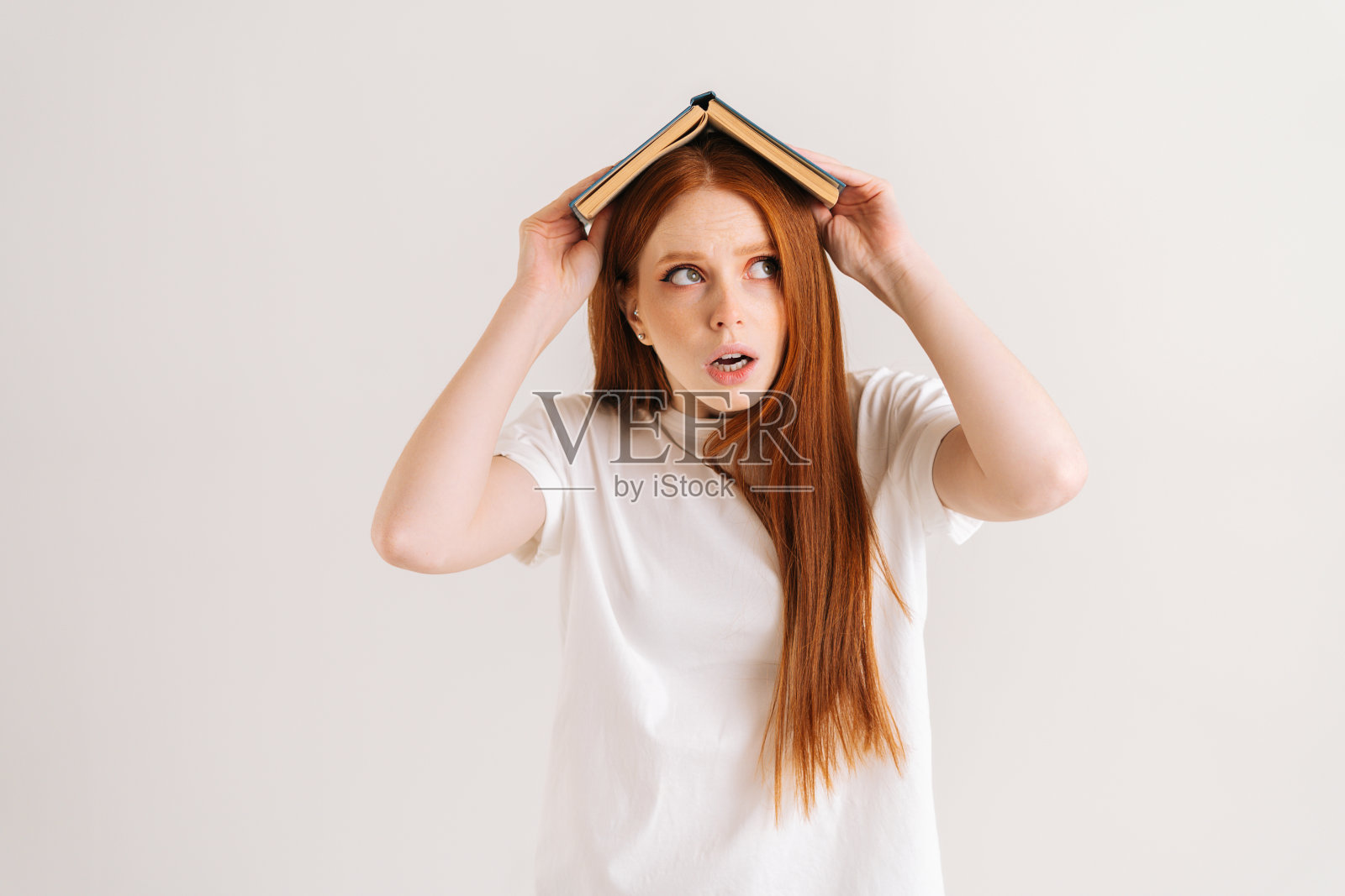 工作室肖像紧张的年轻学生妇女盖着头与书看别处,站在白色孤立的背景。照片摄影图片