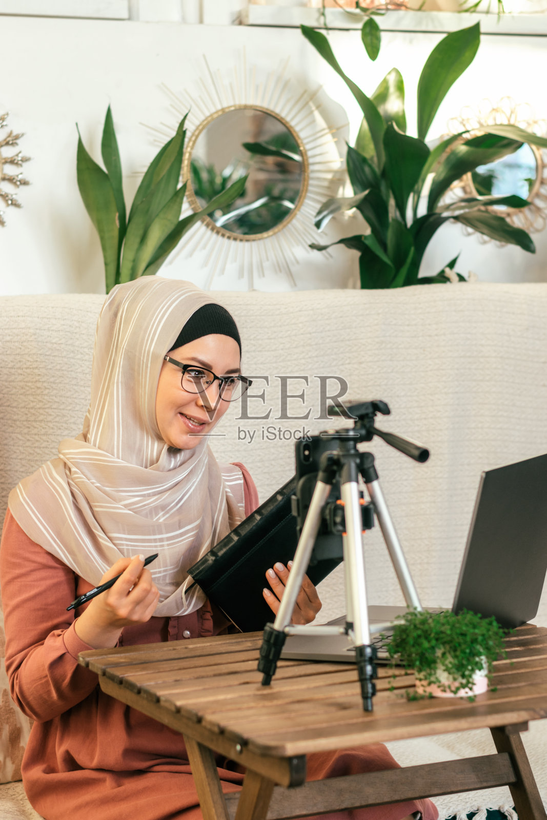 一位戴着头巾的年轻穆斯林妇女主持她的视频博客,并进行现场直播。舒适的室内室内植物。社交媒体和技术概念,多样化的人。照片摄影图片