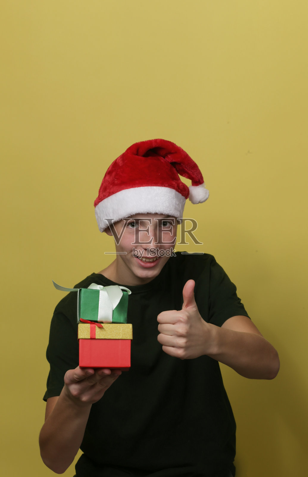 一个十几岁的男孩戴着圣诞老人的帽子,拿着礼物盒,做着拇指的手势。照片摄影图片