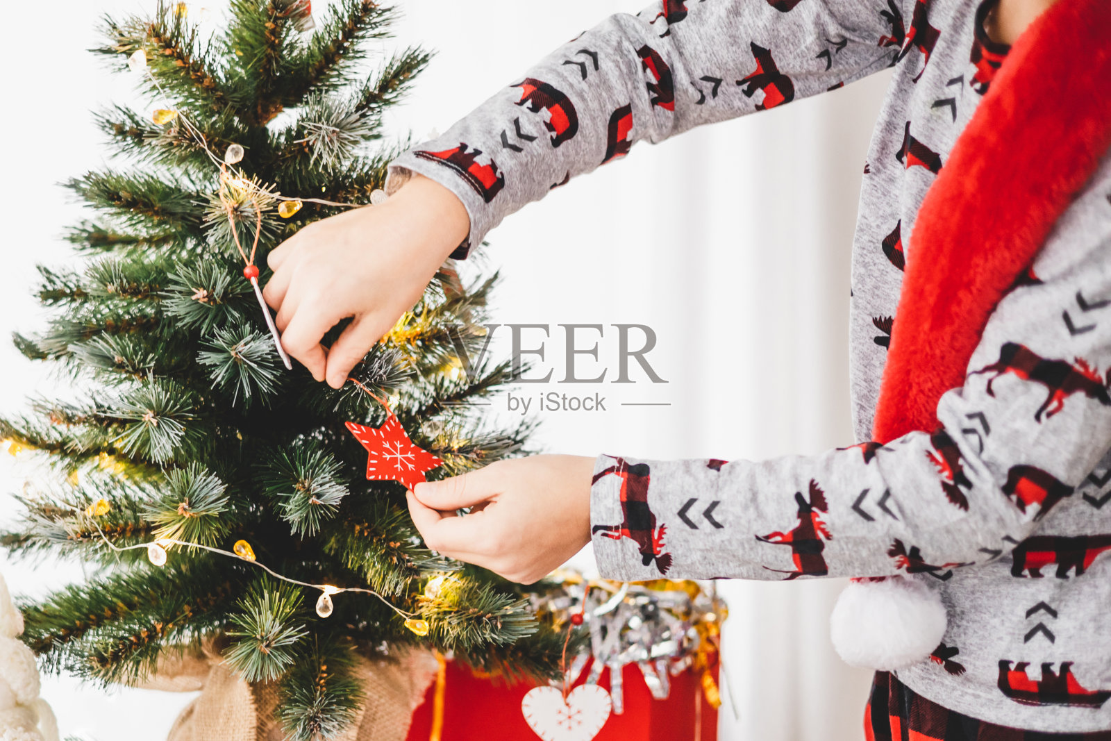 圣诞节和新年假期。小男孩在寒假装饰小圣诞树。穿着睡衣、戴着红色圣诞帽的少年把装饰用的木制玩具挂在冷杉树枝上。寒假照片摄影图片