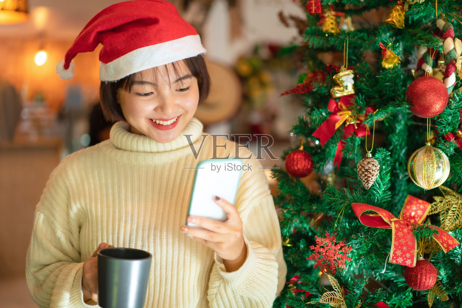 戴圣诞帽的美丽女人手拿咖啡杯,用智能手机与朋友聊天庆祝圣诞节照片摄影图片