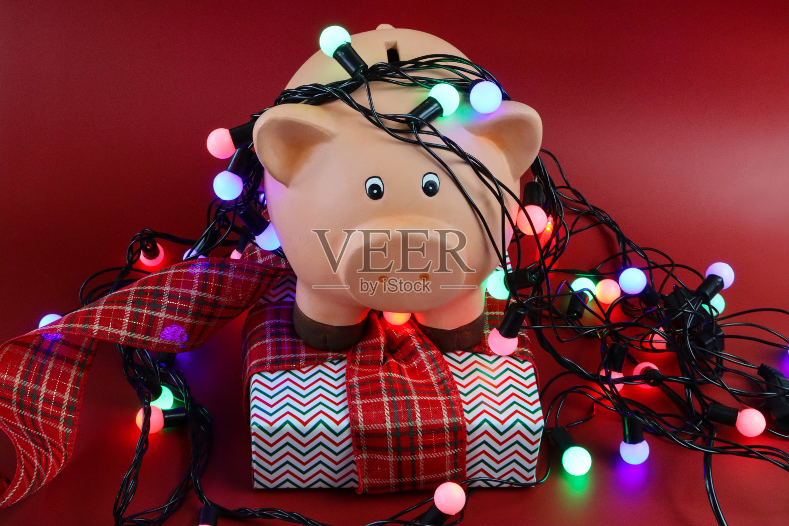 近距离图像的小猪银行包裹着照明的串仙女灯包裹圣诞礼物与格子丝带蝴蝶结,红色的背景,圣诞预算,账单,债务和财务概念照片摄影图片