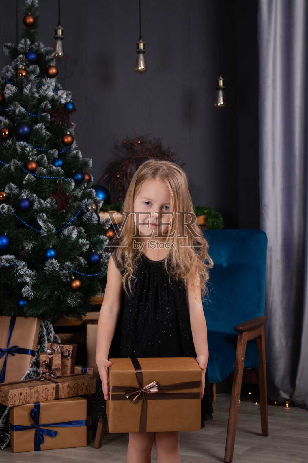 一个可爱的学前班金发小女孩,穿着黑色的衣服,在圣诞树旁拿着新年礼物。黑暗的阁楼房间照片摄影图片