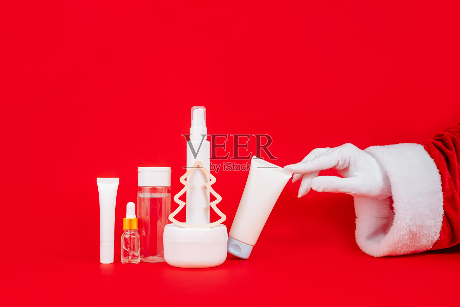 圣诞老人的手在红色背景上触摸日霜、化妆品油、眼霜。没有品牌的瓶子在模型风格。天然化妆品的概念。照片摄影图片