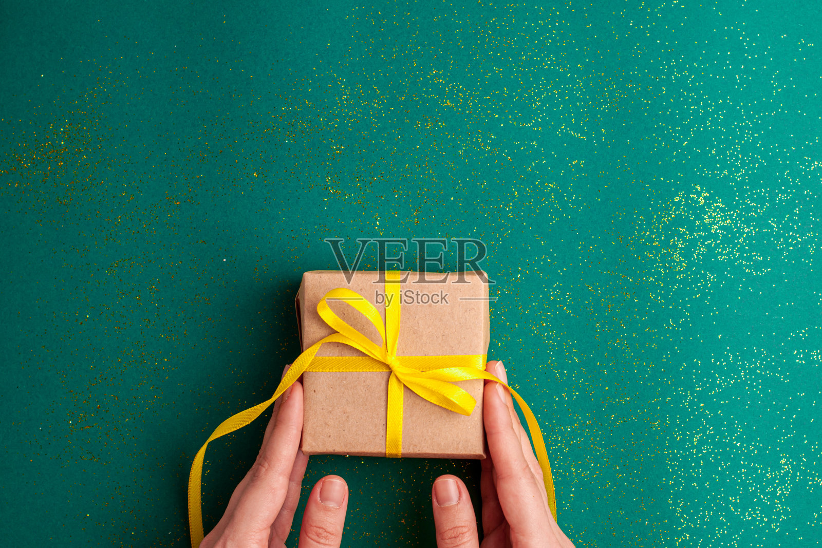 人的双手拿着棕色的礼品盒与一个黄色缎带蝴蝶结在翠绿色的背景与金色的闪光。圣诞假期与现在的概念平铺与复制空间。照片摄影图片