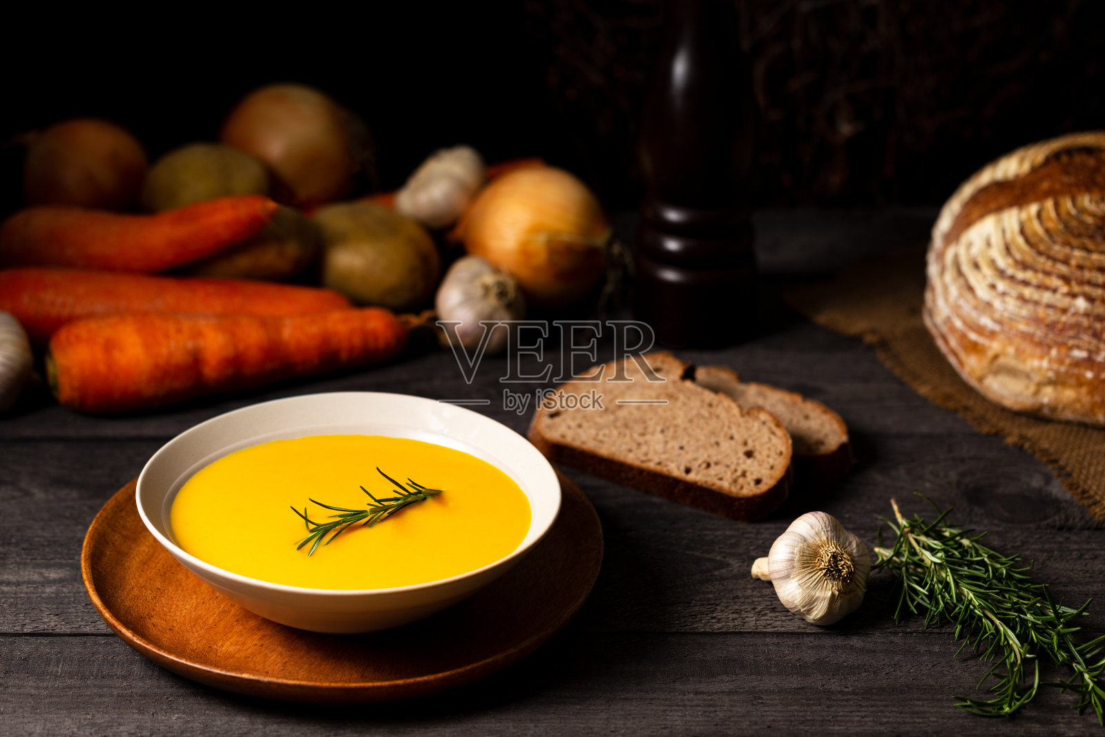 在一张灰色的木头桌子上放上胡萝卜和土豆汤,旁边放上迷迭香枝,还有洋葱,土豆,胡萝卜,大蒜,面包和迷迭香。照片摄影图片