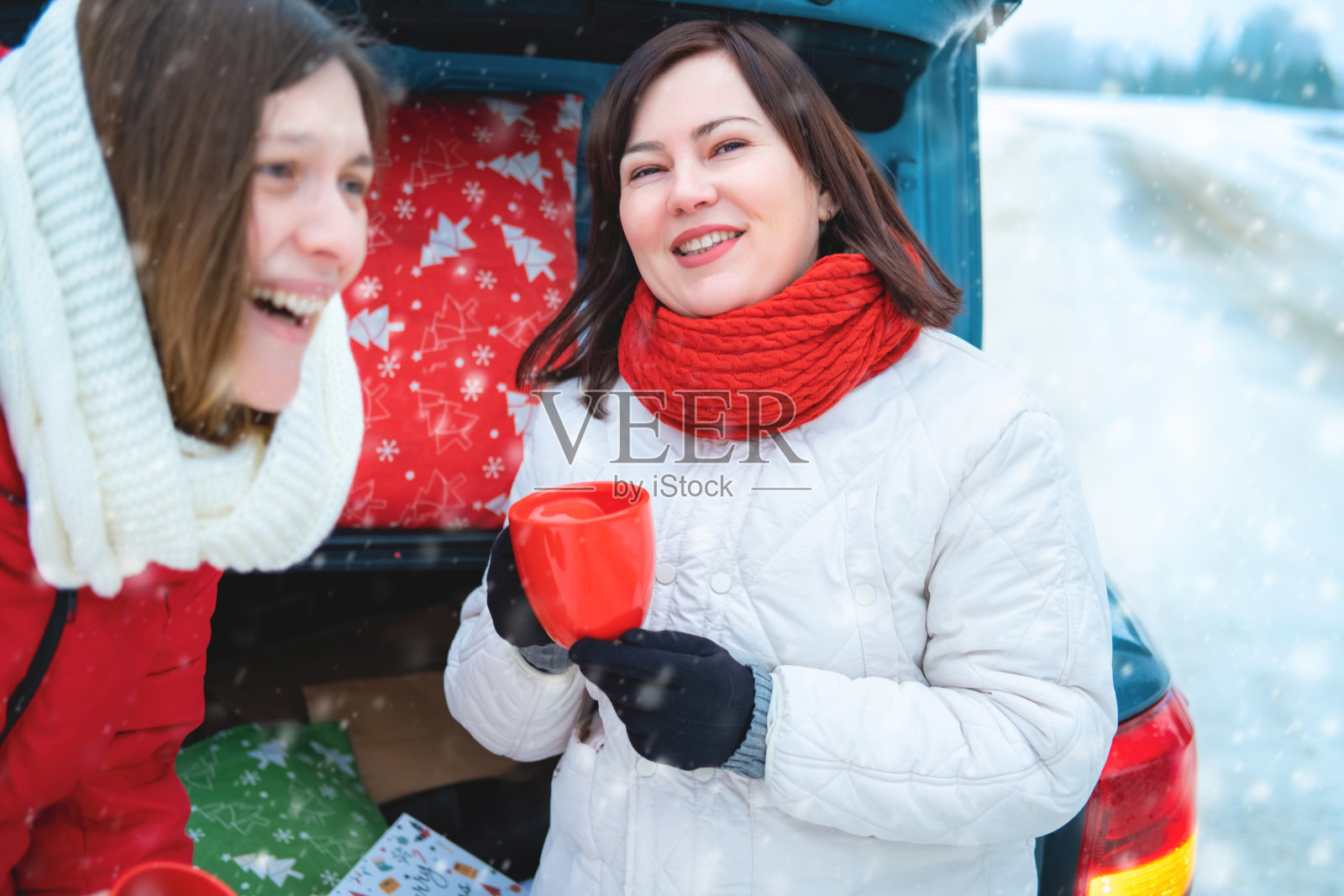 两个年轻的女人在松树林附近的汽车后备箱野餐,在冬季假期开车旅行,喝热巧克力或咖啡,玩得很开心。照片摄影图片