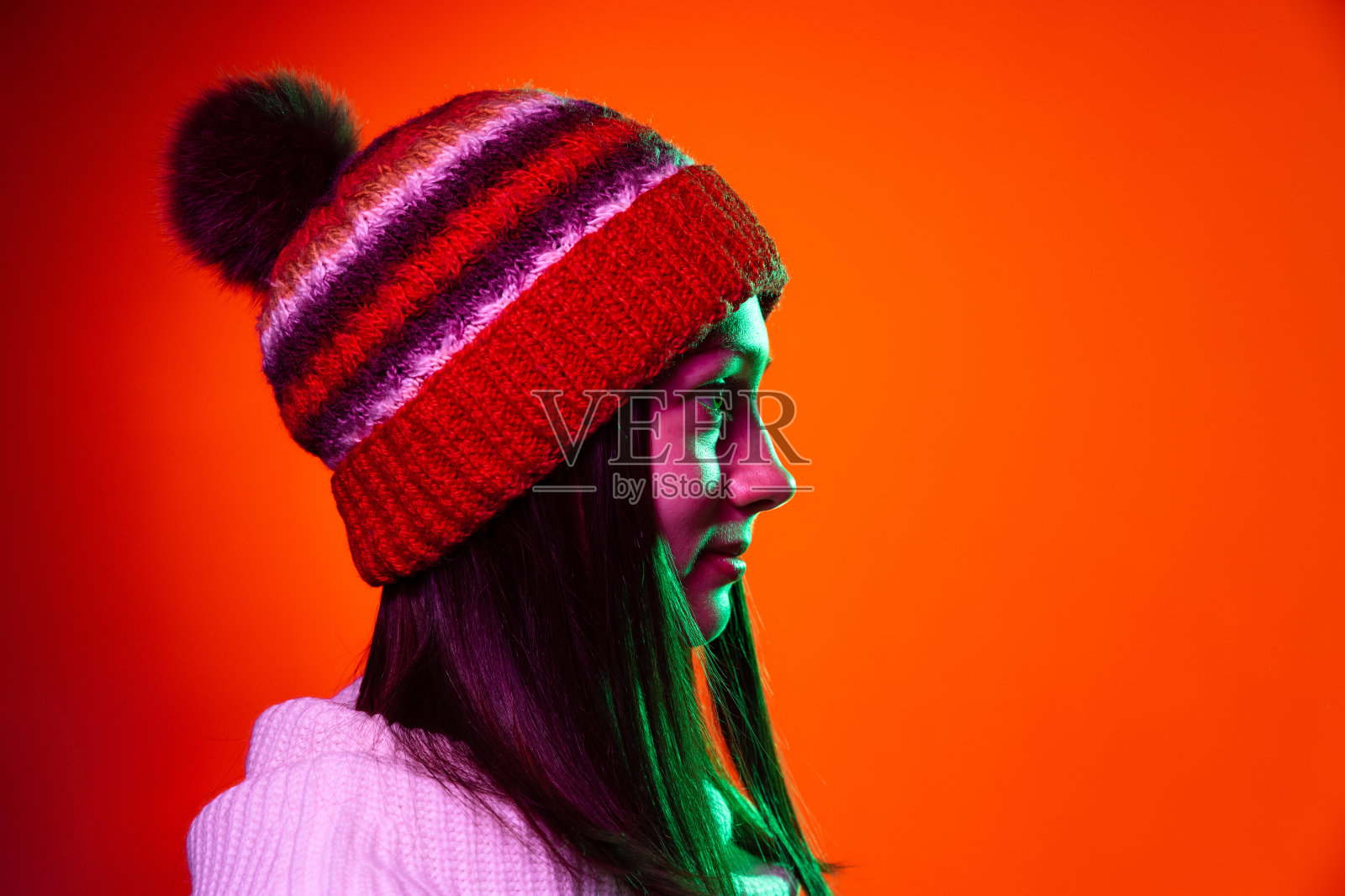 侧面的漂亮的年轻女孩在温暖的针织帽子看着直接孤立在橙色背景霓虹灯。照片摄影图片