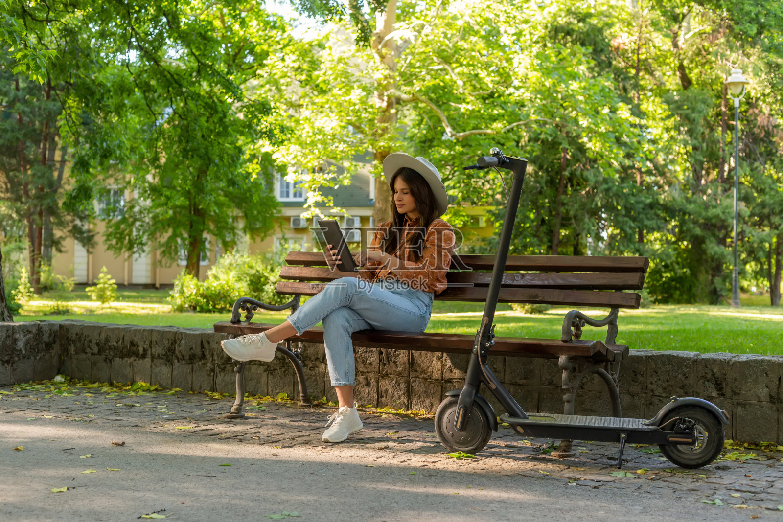 一位戴着帽子的年轻女子坐在公园的长椅上,手里拿着平板电脑滚动着。一辆电动滑板车停在她旁边,背景中以树木和绿色为主。照片摄影图片