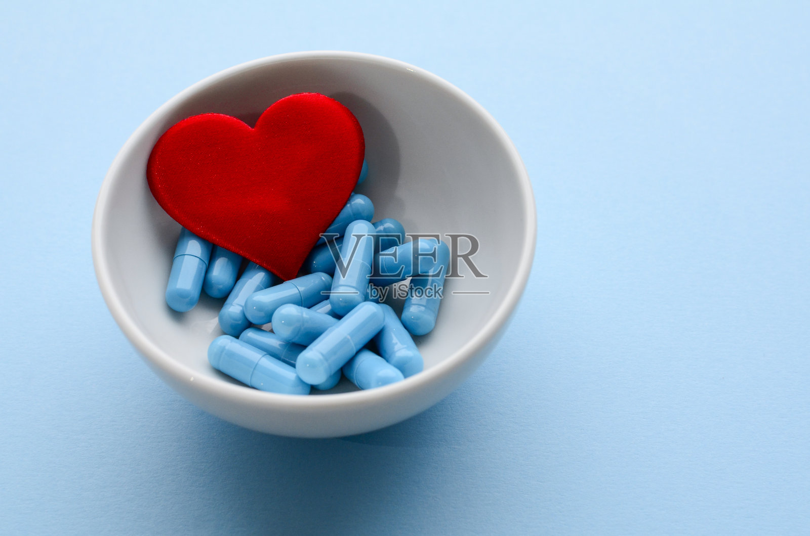 蓝色医药胶囊在白色的碗和红色的心在蓝色的背景。防治心血管疾病,情人节观念。医疗平。照片摄影图片