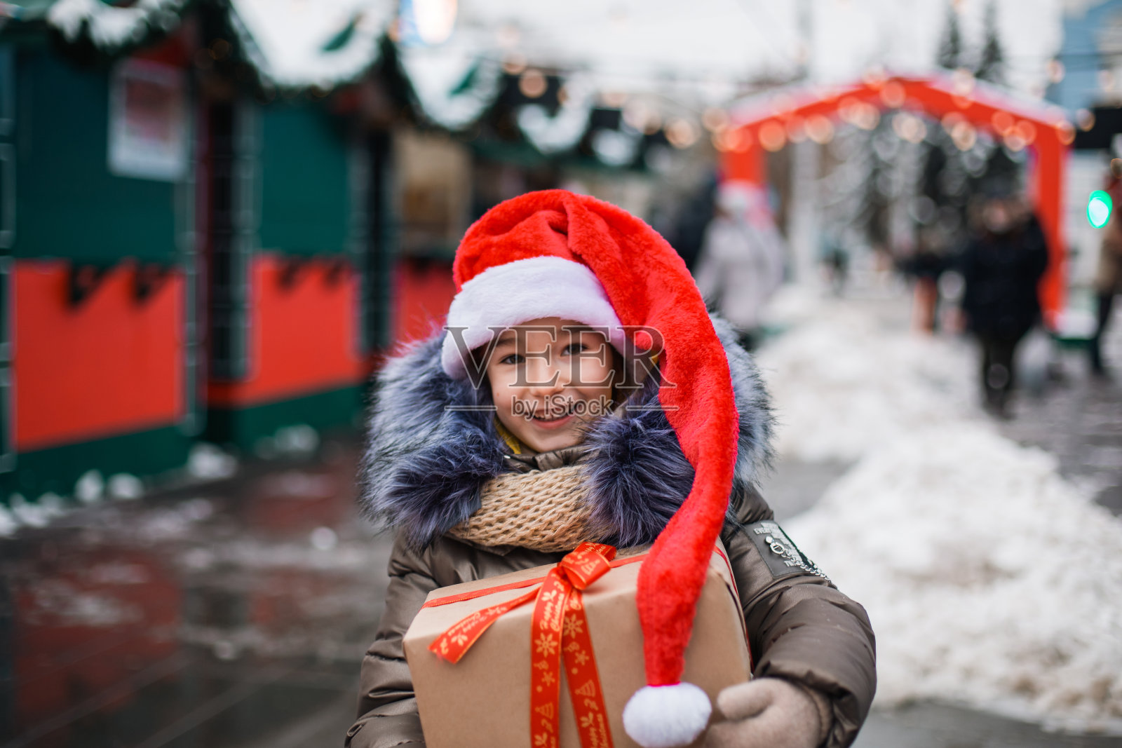 肖像快乐的女孩在圣诞帽与礼物盒的城市街道在冬天的雪在节日市场与装饰和仙女灯。保暖的衣服,针织围巾和毛皮。新的一年照片摄影图片