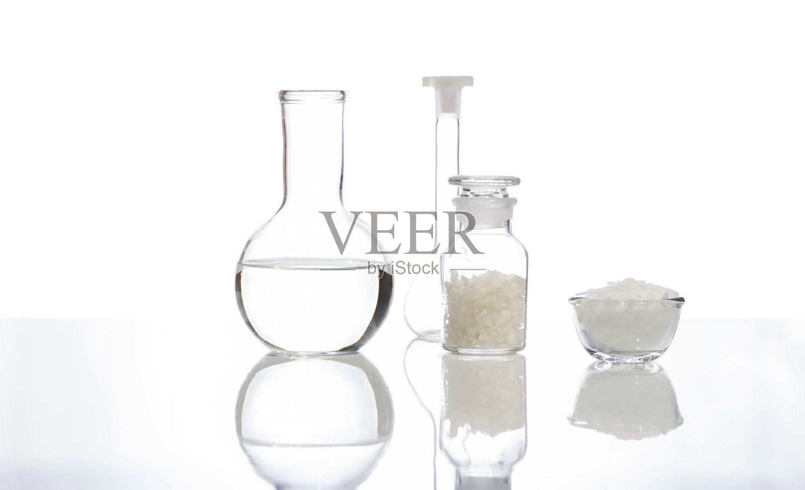 白色片状化学物质在玻璃容器中放置在化学试剂瓶玻璃中的海盐旁边,水晶透明液体在平底烧瓶和容量瓶中。侧视图照片摄影图片