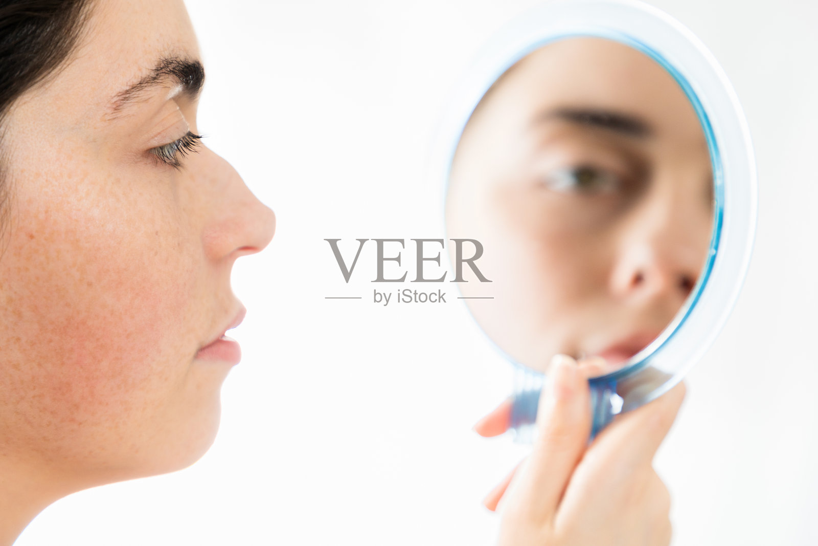 鼻子整形手术。一个年轻的白人妇女的近距离脸看着她的座头鲸鼻子在镜子里,孤立在一个白色的背景。鼻整形术的概念照片摄影图片