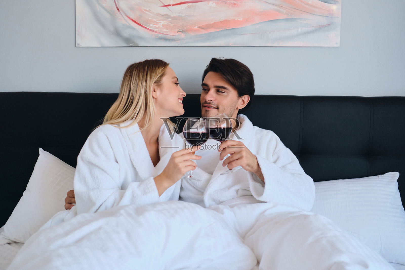 迷人的黑发男人和漂亮的金发女人穿着白色的浴袍,手里拿着一杯红酒,开心地看着对方。年轻漂亮的夫妇躺在现代舒适酒店的床上照片摄影图片