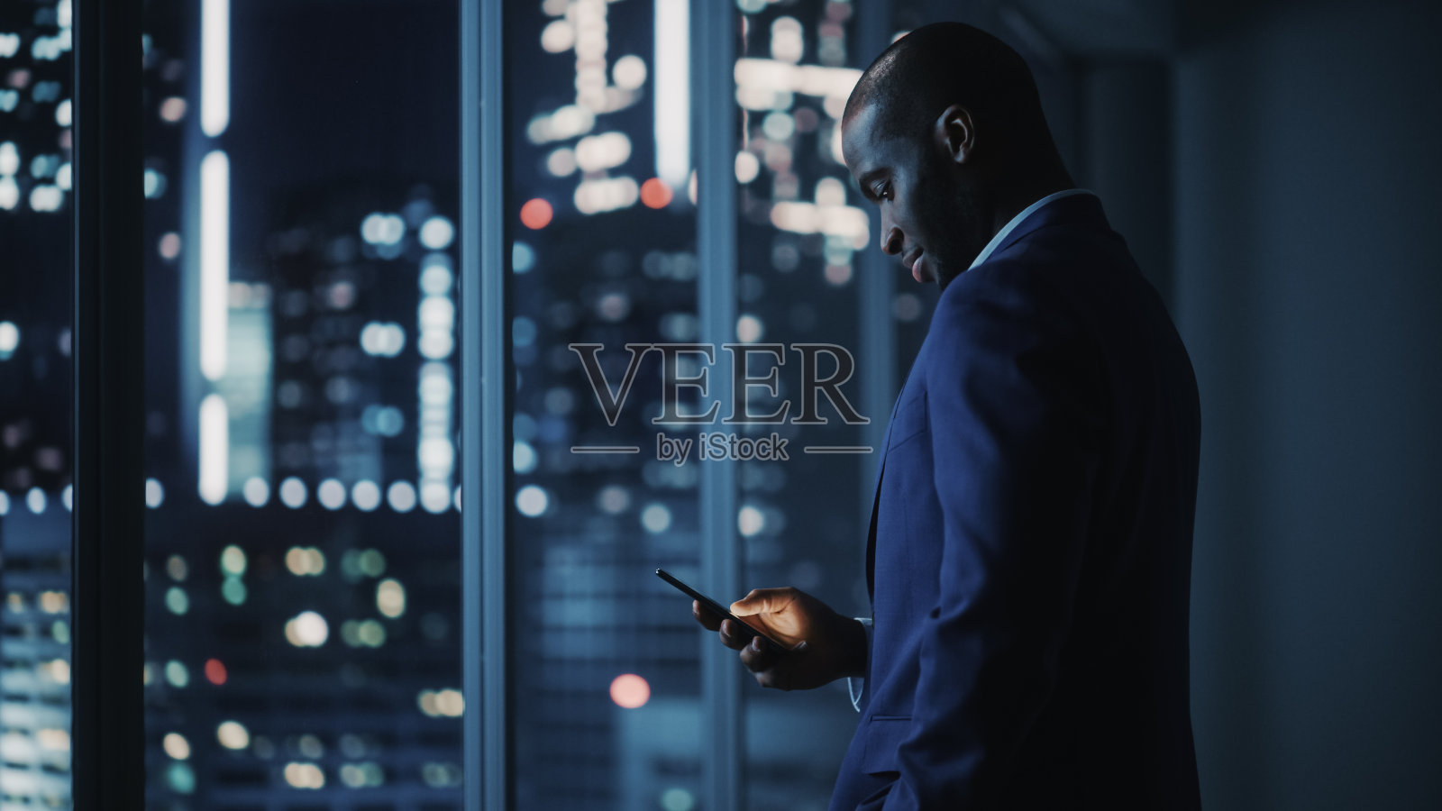 夜间办公室:大城市里穿着西装站着,用智能手机看窗外的成功黑人商人。非裔美国CEO对电子商务软件投资策略的思考照片摄影图片