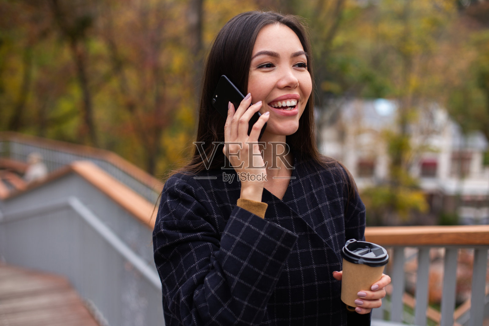 年轻积极随意的女人,穿着外套,喝着咖啡,兴高采烈地打着手机,在公园里散步照片摄影图片