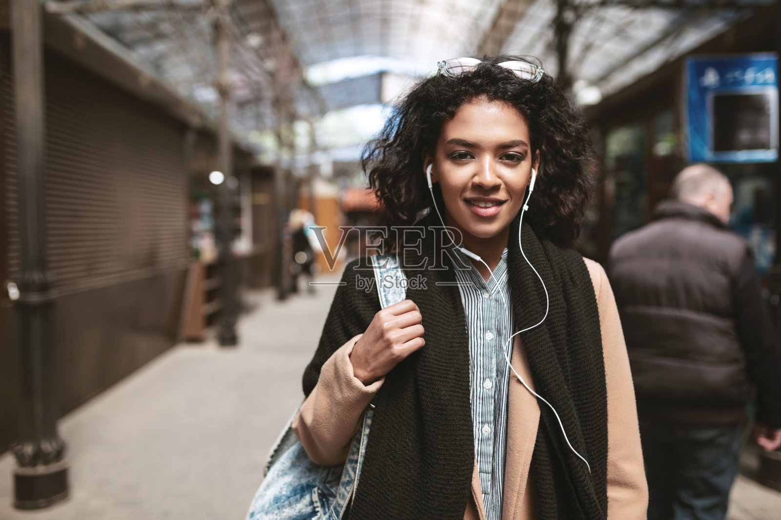 漂亮的非洲裔美国女孩的肖像站在街上,看着在相机里酷女孩深色卷发在耳机里走在城市的外套,围巾和包在她的肩膀上照片摄影图片