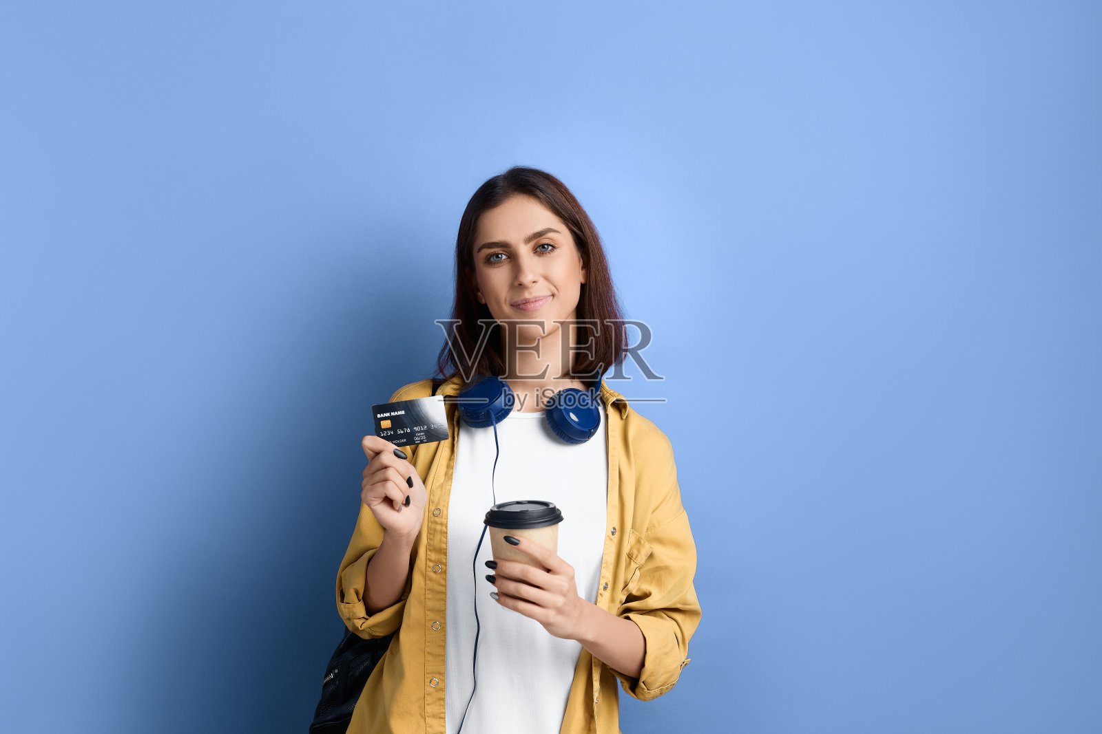 年轻的女学生拿着外卖咖啡,出示信用卡,很高兴使用银行服务和在线支付,穿着黄色的衬衫,白色的t恤,黑色的包,脖子上戴着耳机。照片摄影图片