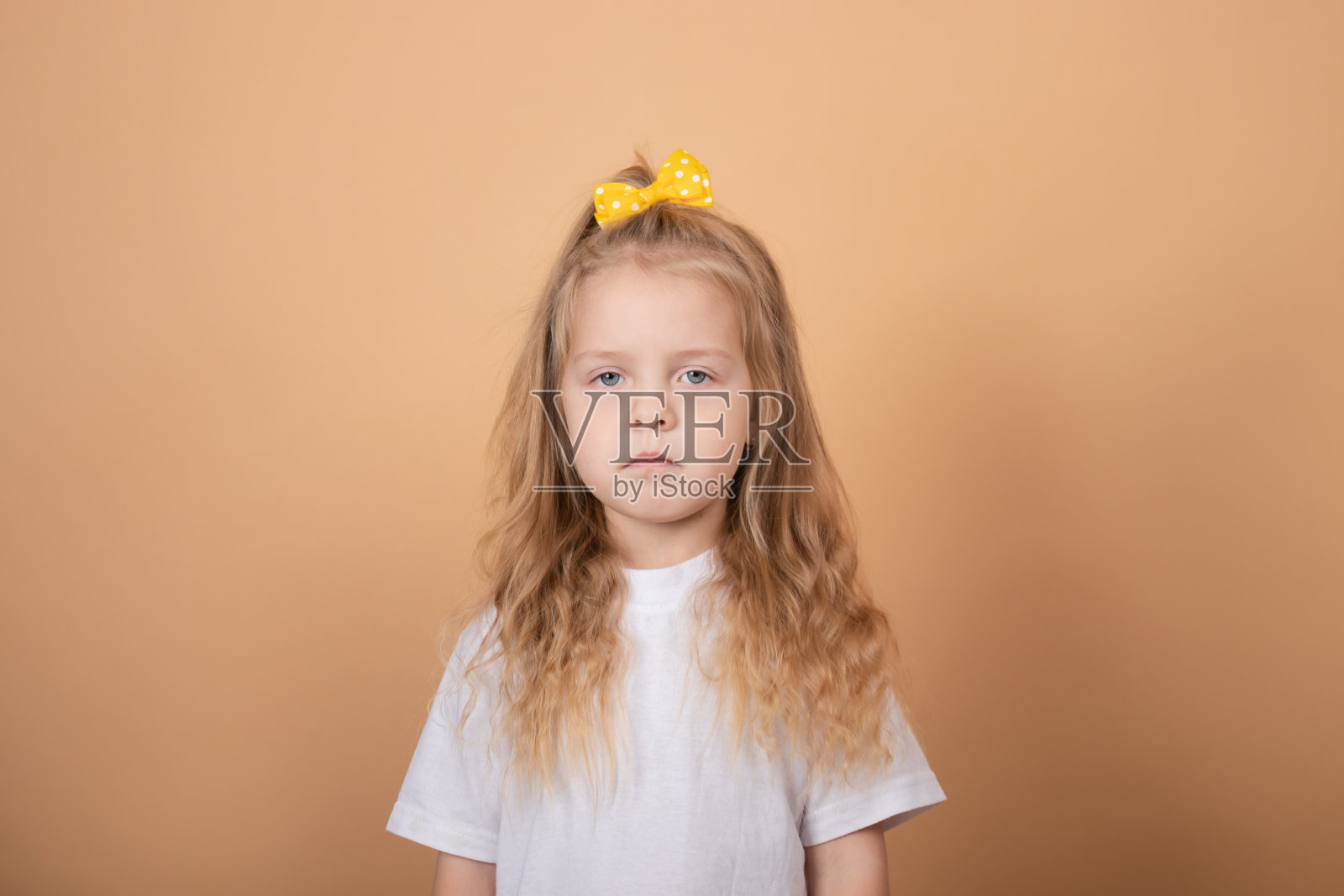 一个可爱的金发小女孩的肖像,穿着白色的t恤,头上戴着黄色的蝴蝶结。棕色的背景照片摄影图片