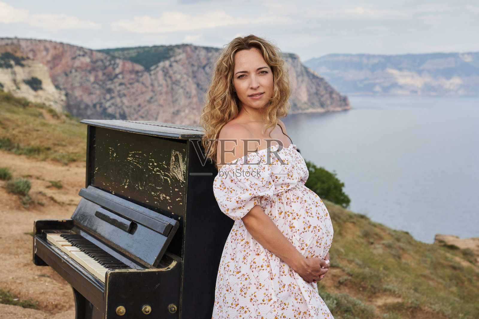 老钢琴在田野里,在岩石海岸的背景上。年轻的孕妇靠着他站着照片摄影图片