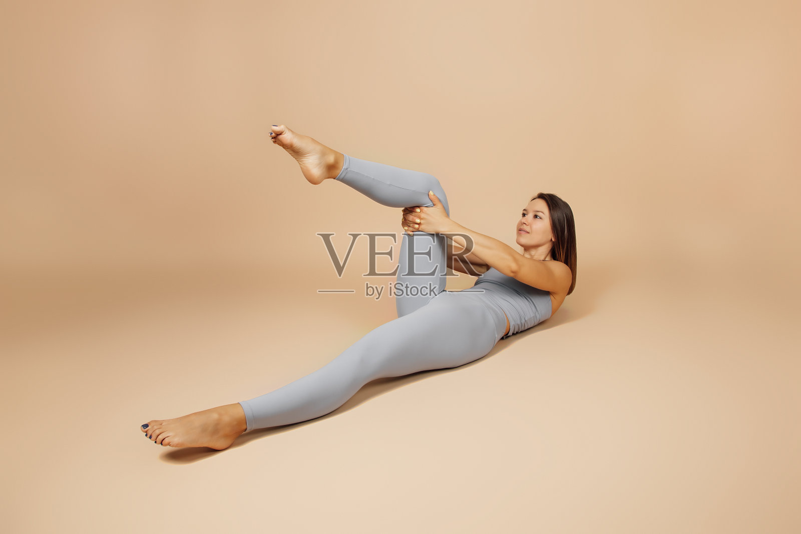 布鲁内特热衷于健身。美丽的年轻女子在灰色紧身瑜伽服做运动在中性的米色背景下,她躺在她的背部,她的膝盖到她的胸部,侧视照片摄影图片