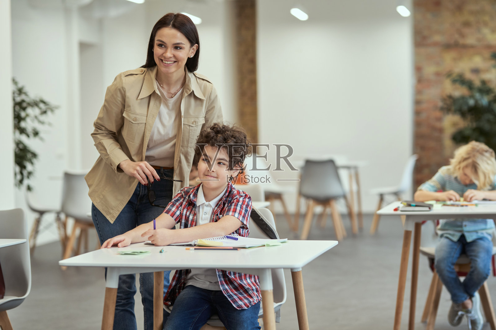 鼓励孩子学习。在课堂上,年轻的女老师面带微笑地站在小男生旁边。孩子们在上小学,坐在课桌前照片摄影图片