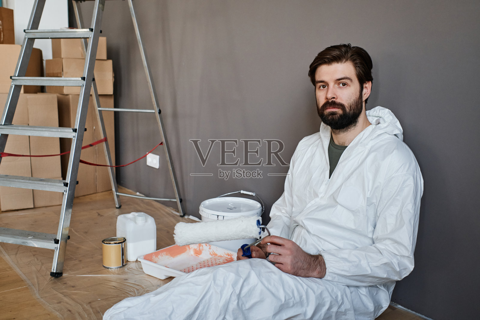 年轻的白人男子穿着防护服在新公寓里做装修工作,坐在地板上看着摄像机照片摄影图片