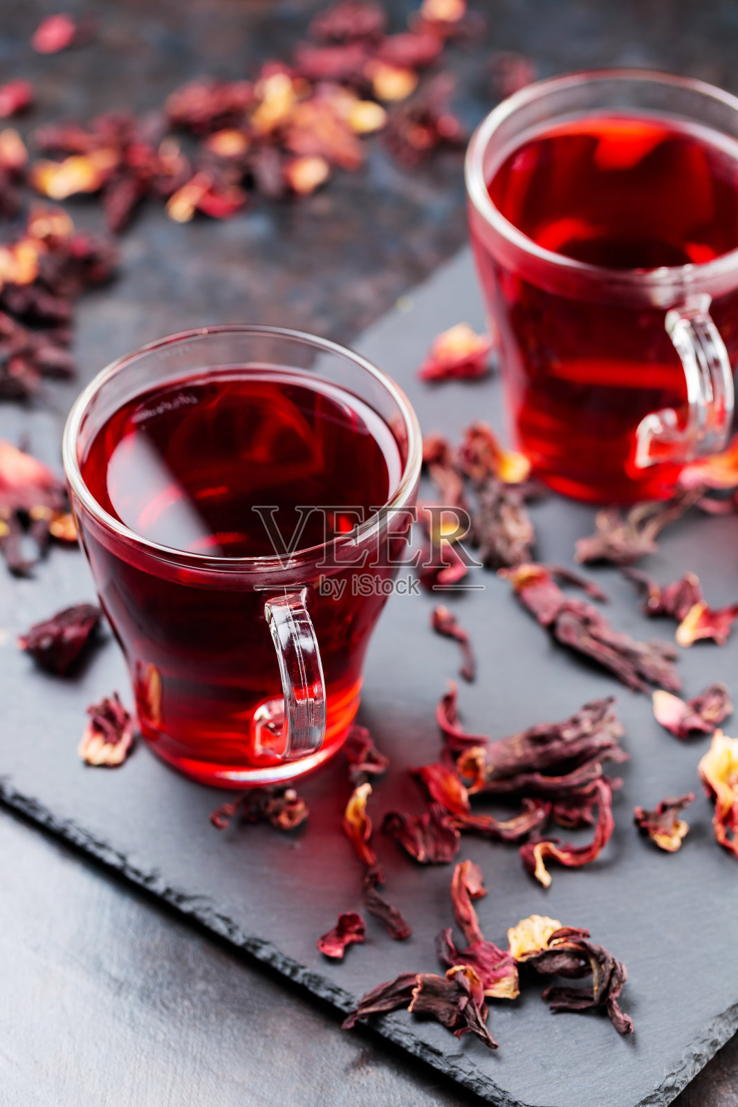 黑色背景的木槿茶玻璃杯。一杯红木槿茶和干木槿花瓣放在石板上照片摄影图片