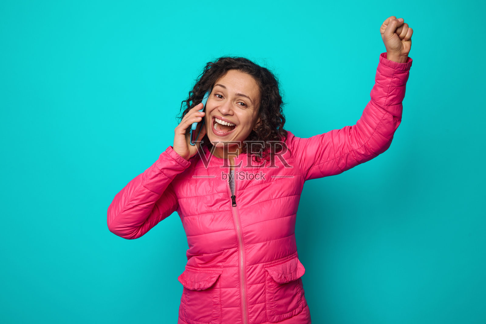 穿着亮粉色夹克的棕发年轻女孩,双手紧握,一边说着手机,一边看着镜头,表达着快乐与喜悦。孤立在蓝色背景,复制广告空间照片摄影图片