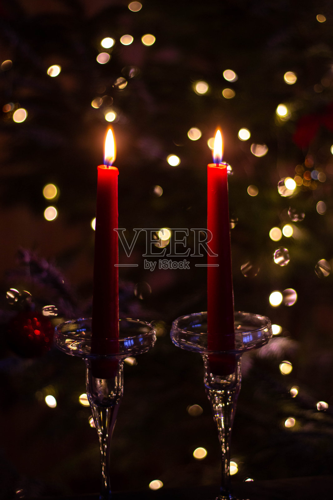 两支红色长蜡烛插在高高的玻璃烛台上,背景是新年树的灯光。烛光浪漫的新年。背景的散景效果照片摄影图片