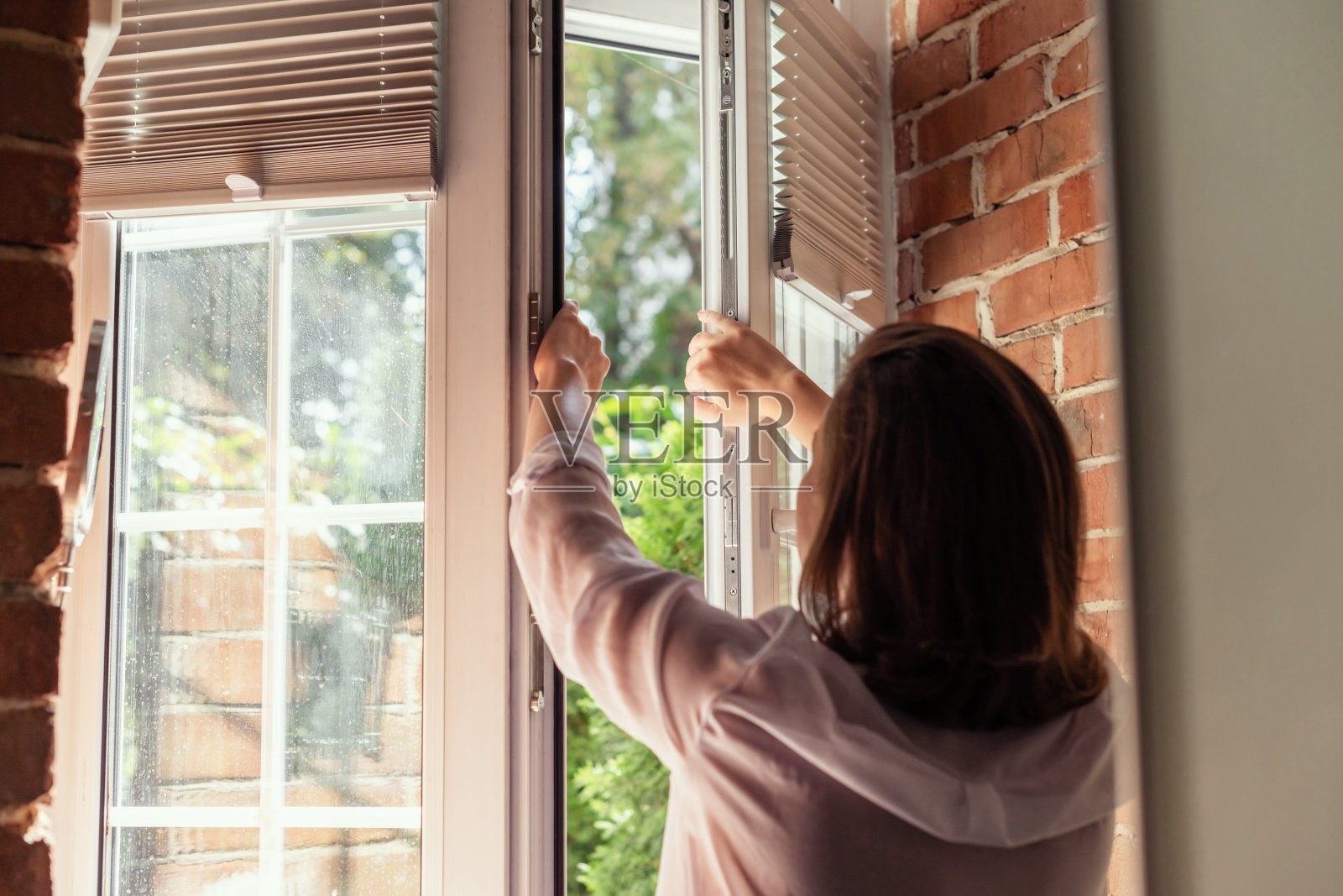 身着睡衣的年轻美女在初夏的清晨打开乡村小屋的房间窗户,呼吸着新鲜空气。后视图。照片摄影图片