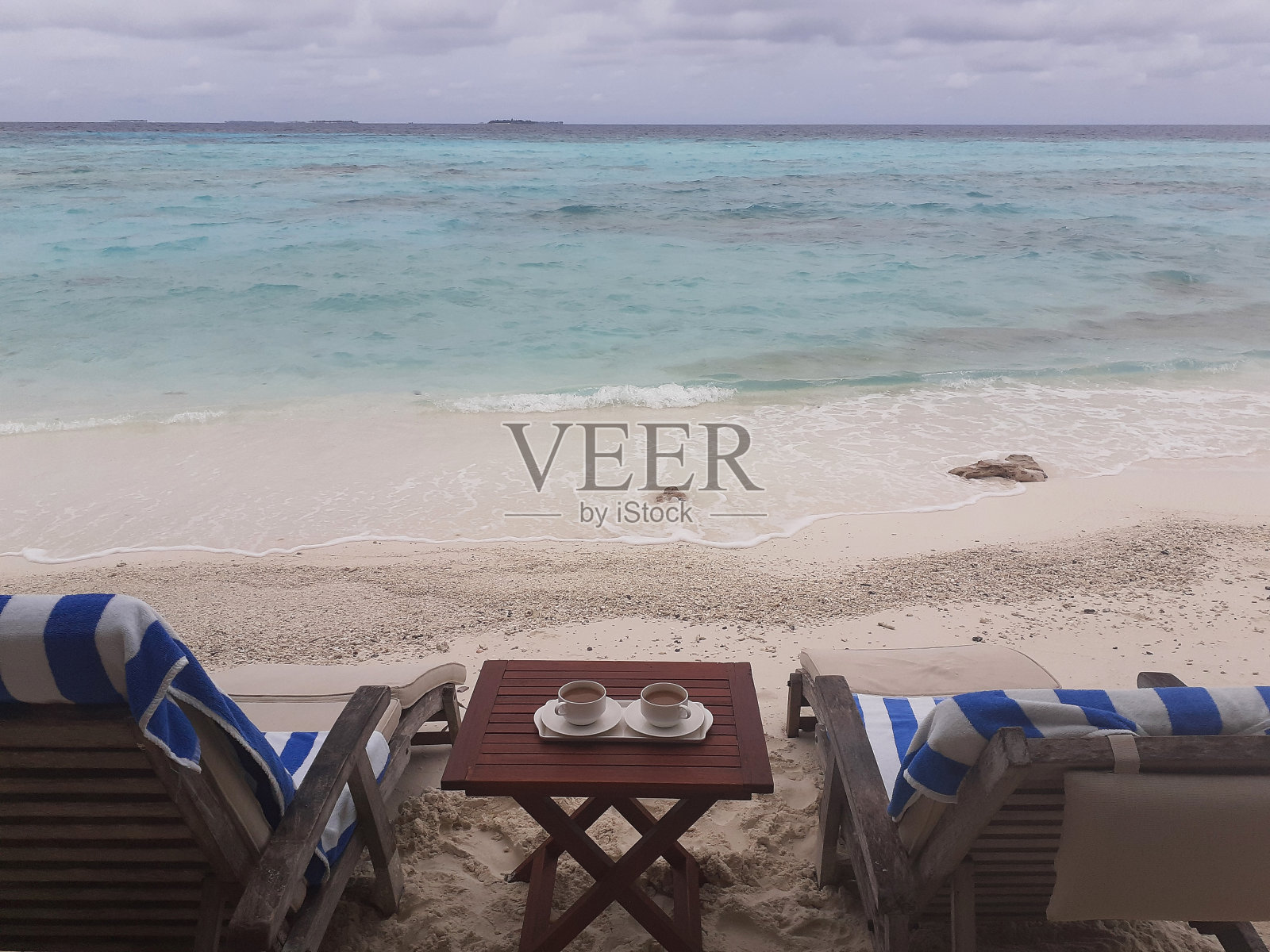 两张空的日光浴床和木桌上的两杯咖啡。马尔代夫,靠近蓝绿色海洋的白色沙滩。照片摄影图片