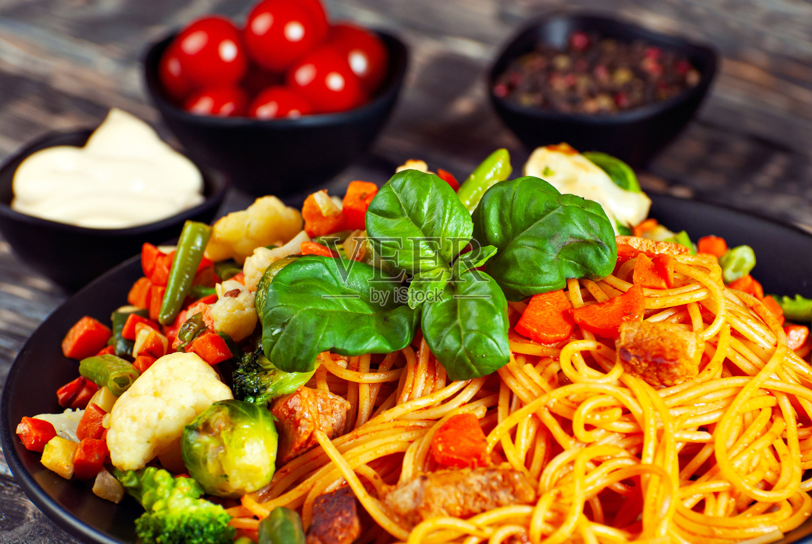 黑色盘子里有胡萝卜、蔬菜和肉的面条。黑色的埃里温餐桌上的食物。罗勒叶在意大利面上。混合蔬菜的意大利面。背景是腌番茄和香料。照片摄影图片