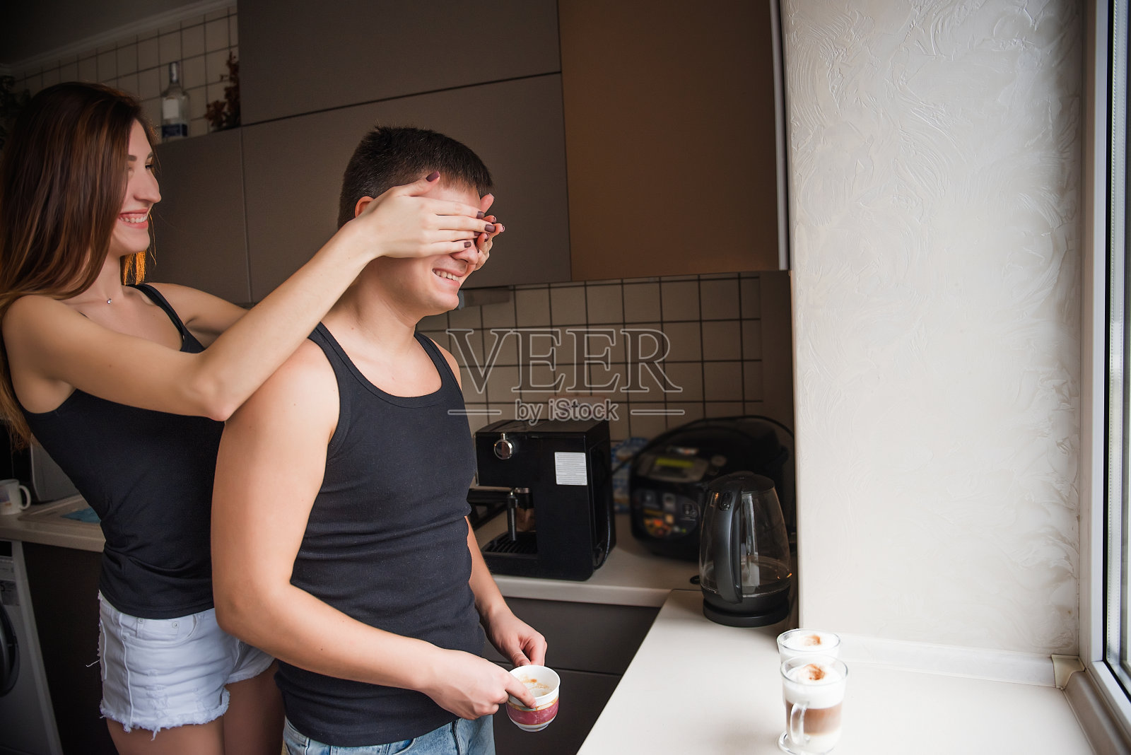 女人对窗边的男人闭眼。一对千禧一代的年轻夫妇在厨房里拥抱照片摄影图片