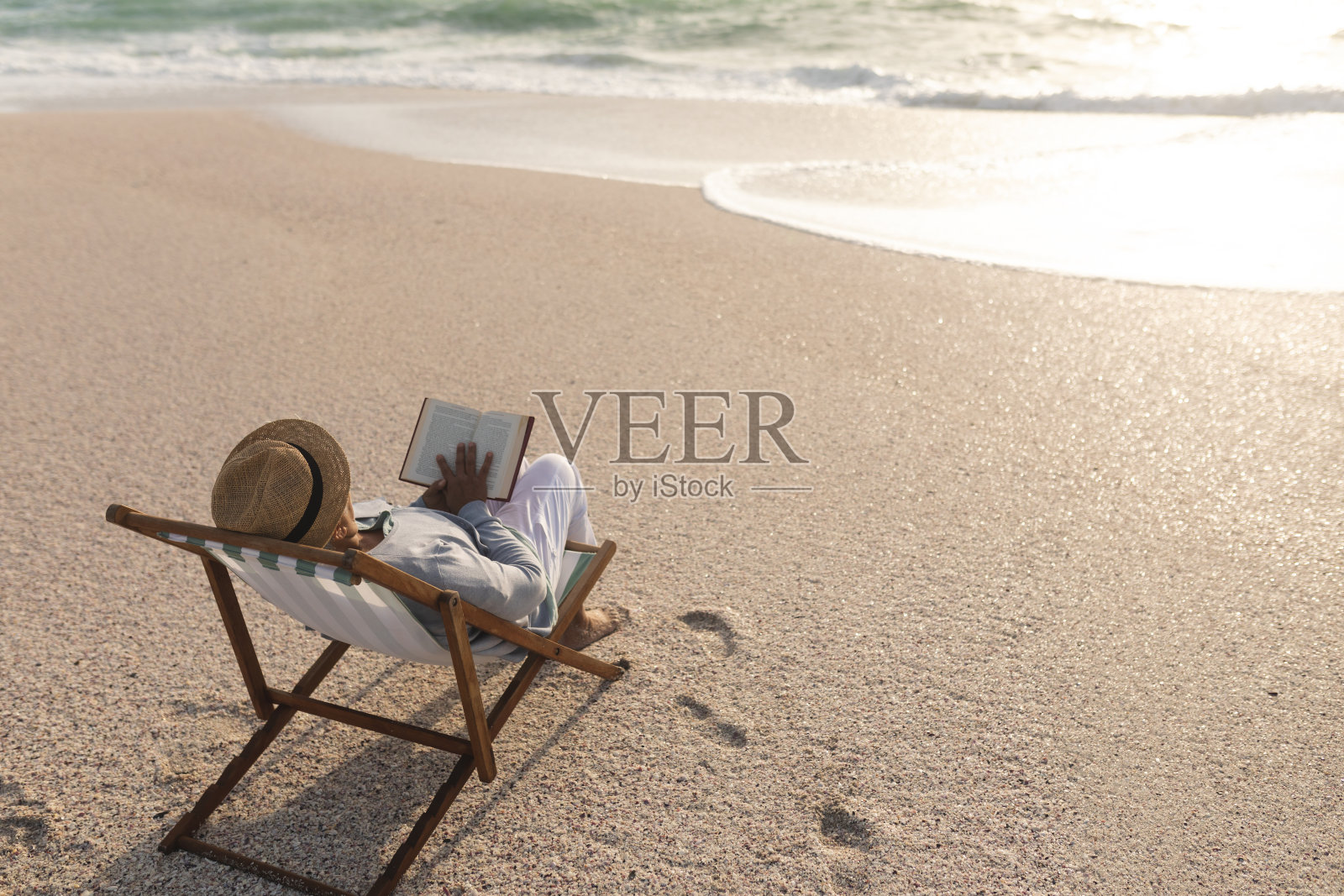 放松的年长混血儿妇女阅读的书,而坐在折叠椅在海滩在日落照片摄影图片