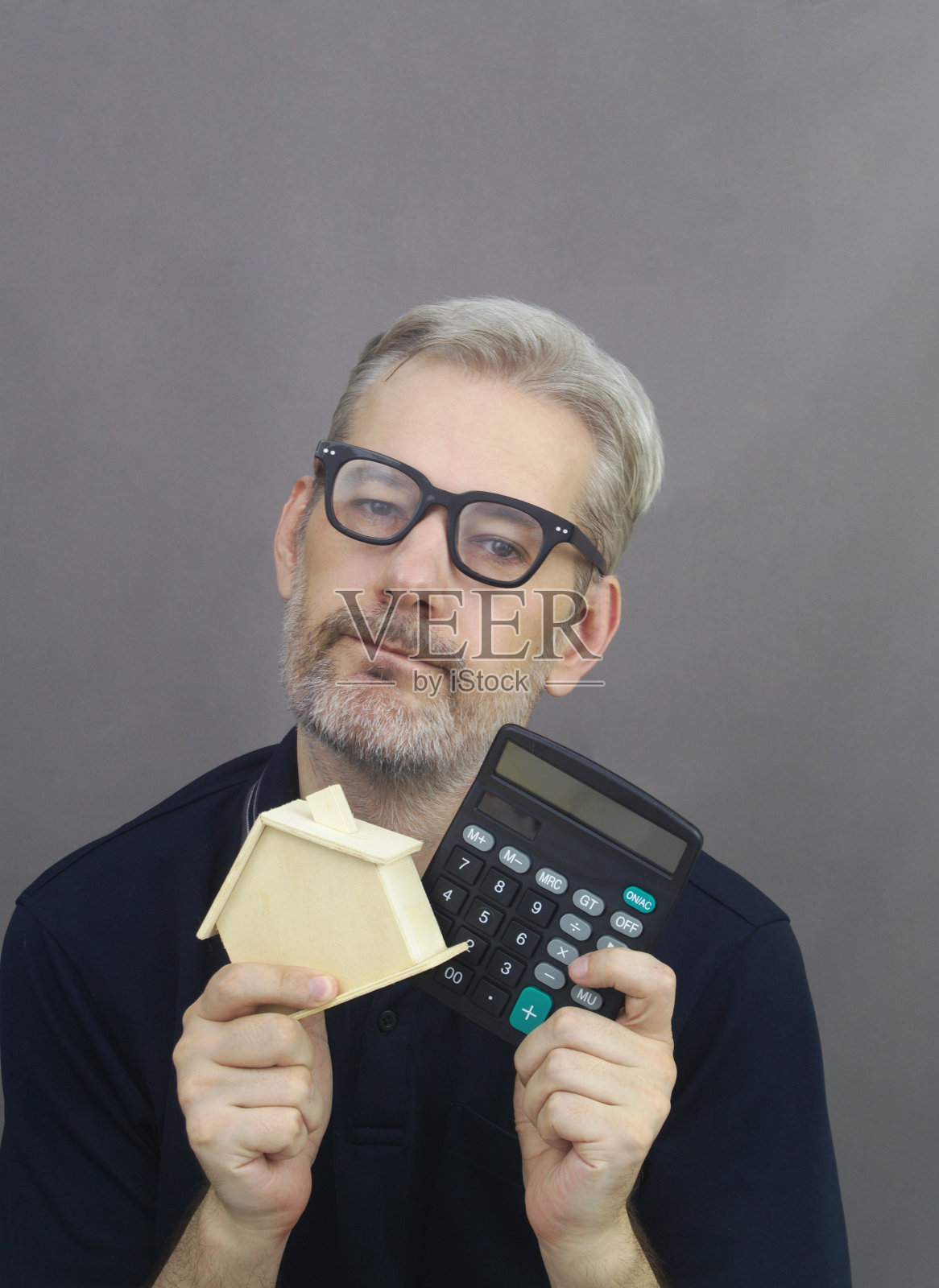 聪明的中年男人,灰色的头发和胡子,拿着木制房子模型和计算器在灰色的背景。照片摄影图片
