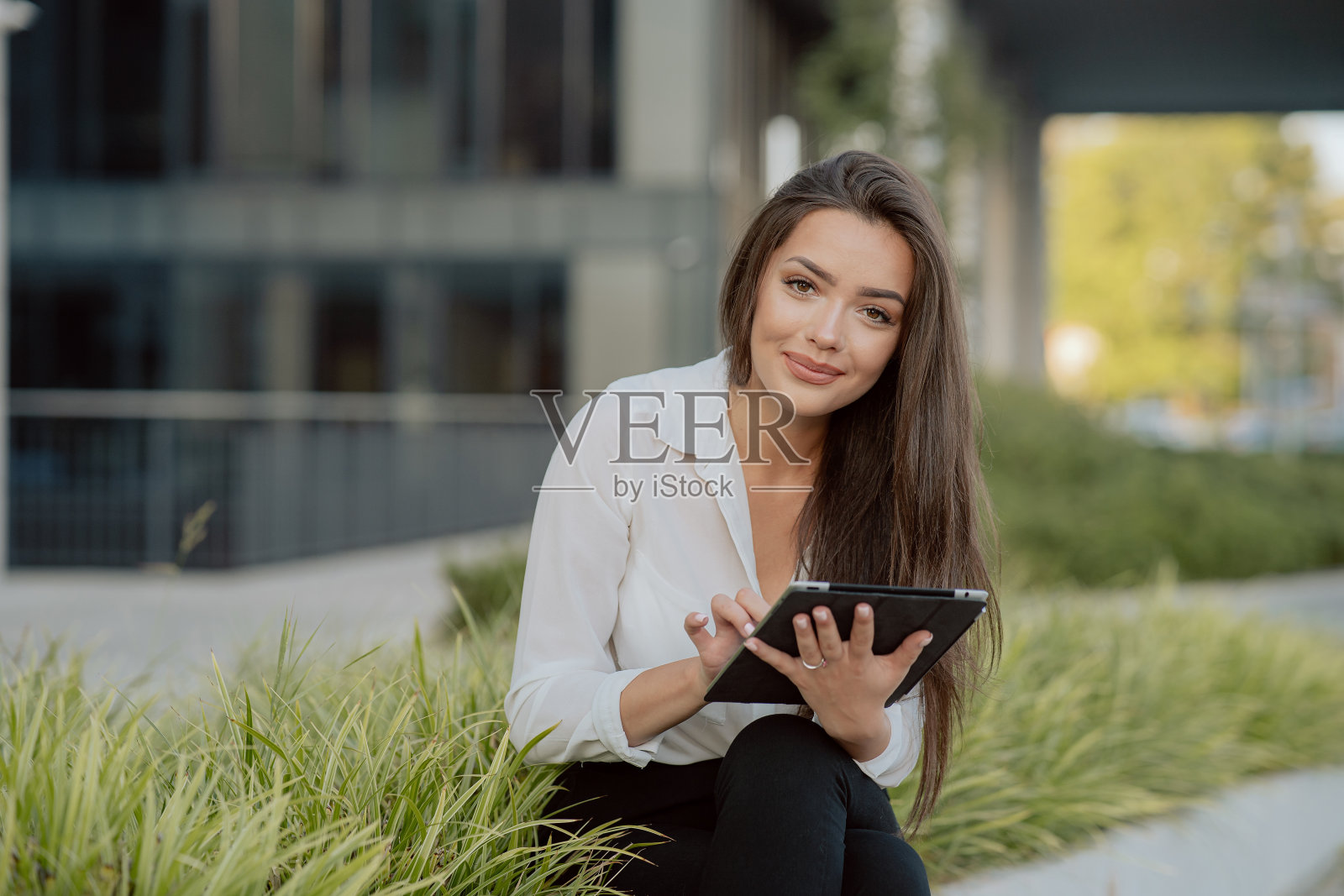 离开办公室,穿着漂亮的女孩坐在现代企业大楼前的混凝土上,周围是观赏植物,一个女人拿着平板电脑,上网,浏览社交媒体照片摄影图片