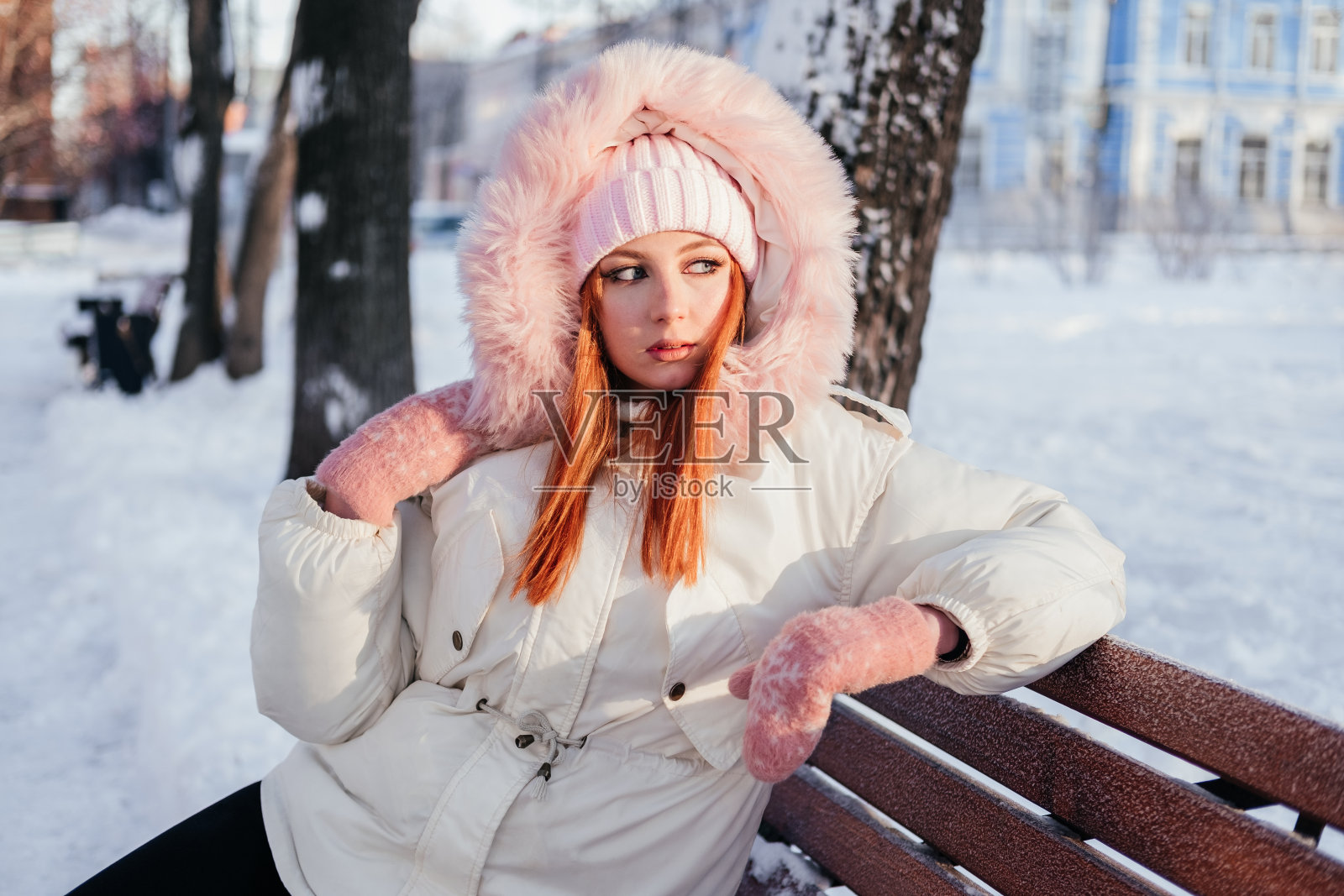 一个穿着外套、戴着手套、戴着帽子的姜黄色年轻女子坐在冬季公园户外的长椅上。城市街道上的红发女孩照片摄影图片