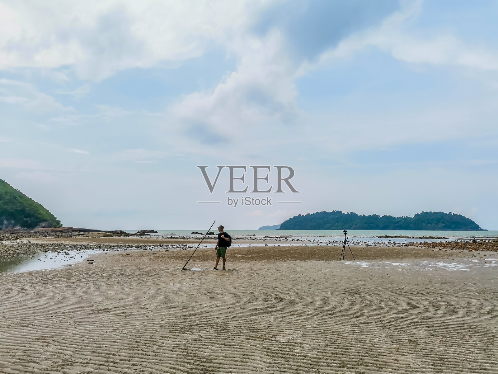 男性背包客站在低潮的沙滩上与岛屿和多云的天空背景。寻找灵感的摄影师。沙滩上的三脚架。旅行生活冒险概念照片摄影图片