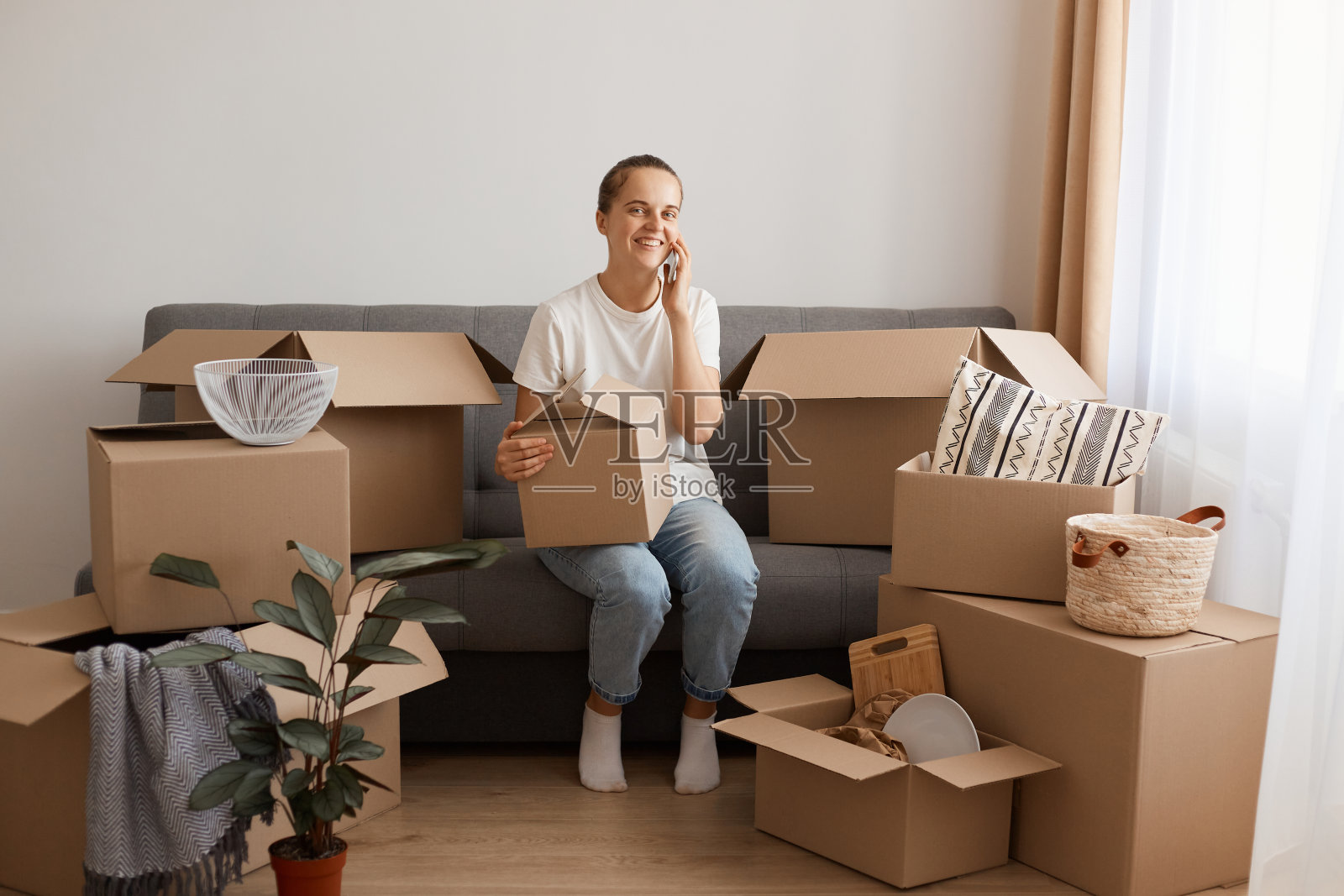 一个穿着白色t恤和牛仔裤微笑着咳嗽的女人,在搬家后的新公寓里,拿着纸板箱和电话摆姿势,吹嘘她的公寓,谈论着搬家的事。照片摄影图片