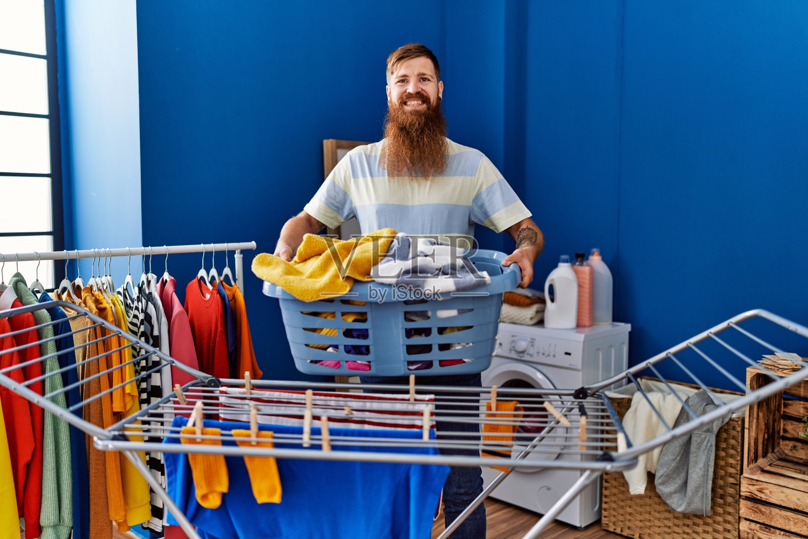 年轻的红发男子微笑着自信地拿着篮子衣服在洗衣房照片摄影图片