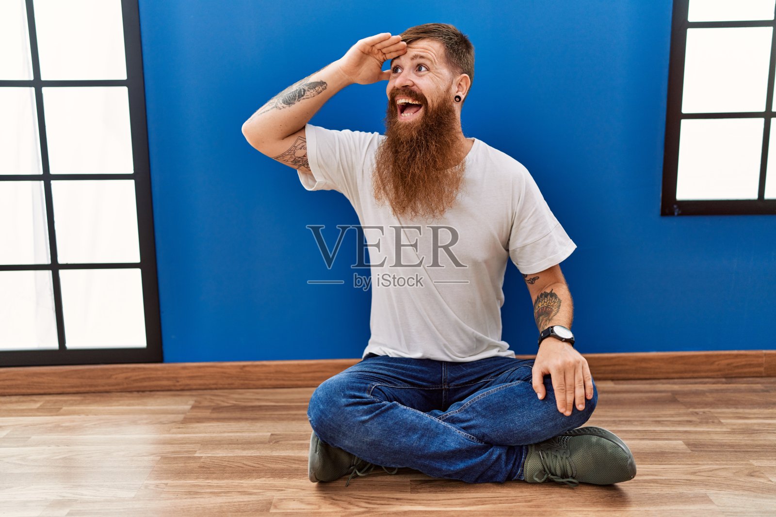 一个留着长胡子的红头发男人坐在空房间的地板上,非常高兴,微笑着,双手举过头顶望着远方。搜索概念。照片摄影图片