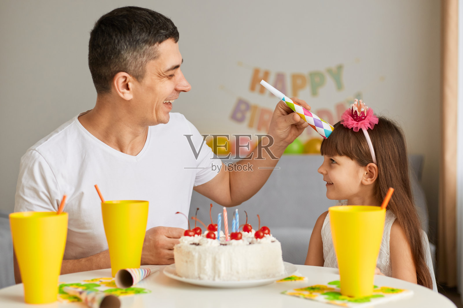 穿着白色t恤的英俊男子为女儿庆祝生日,用派对喇叭玩得很开心,坐在有蜡烛的节日蛋糕的桌子旁。照片摄影图片