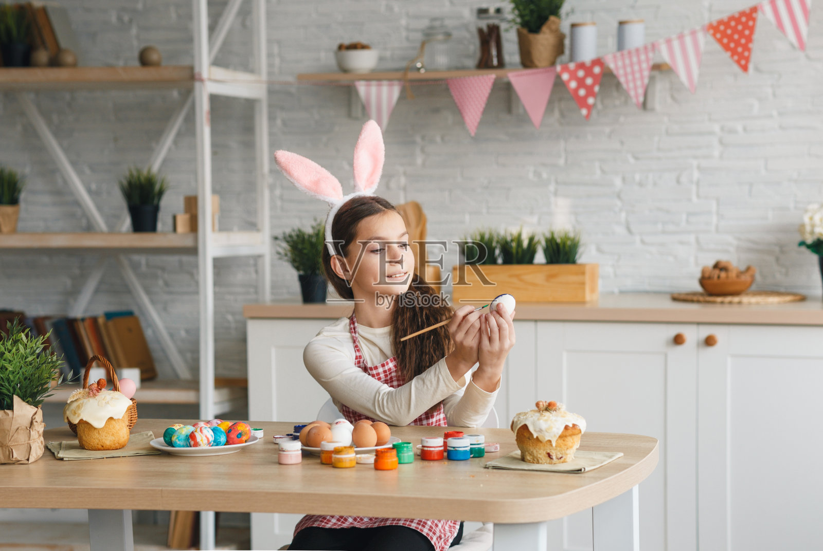 一个戴着兔耳朵的可爱小女孩正坐在厨房的桌子旁准备复活节彩蛋。照片摄影图片