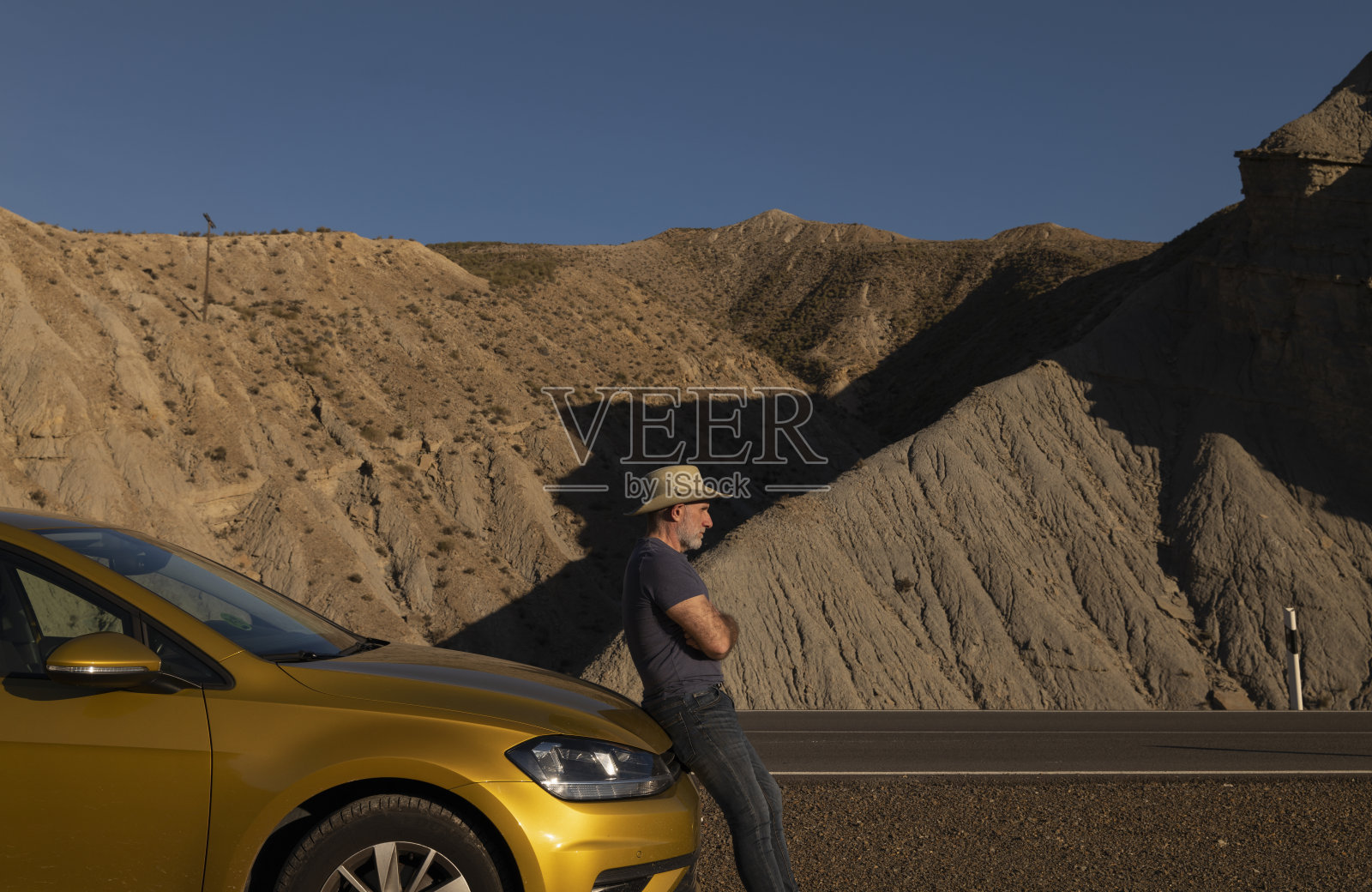 一个戴着牛仔帽的成年男子站在汽车前,在沙漠中靠山而行。西班牙阿尔梅里亚照片摄影图片