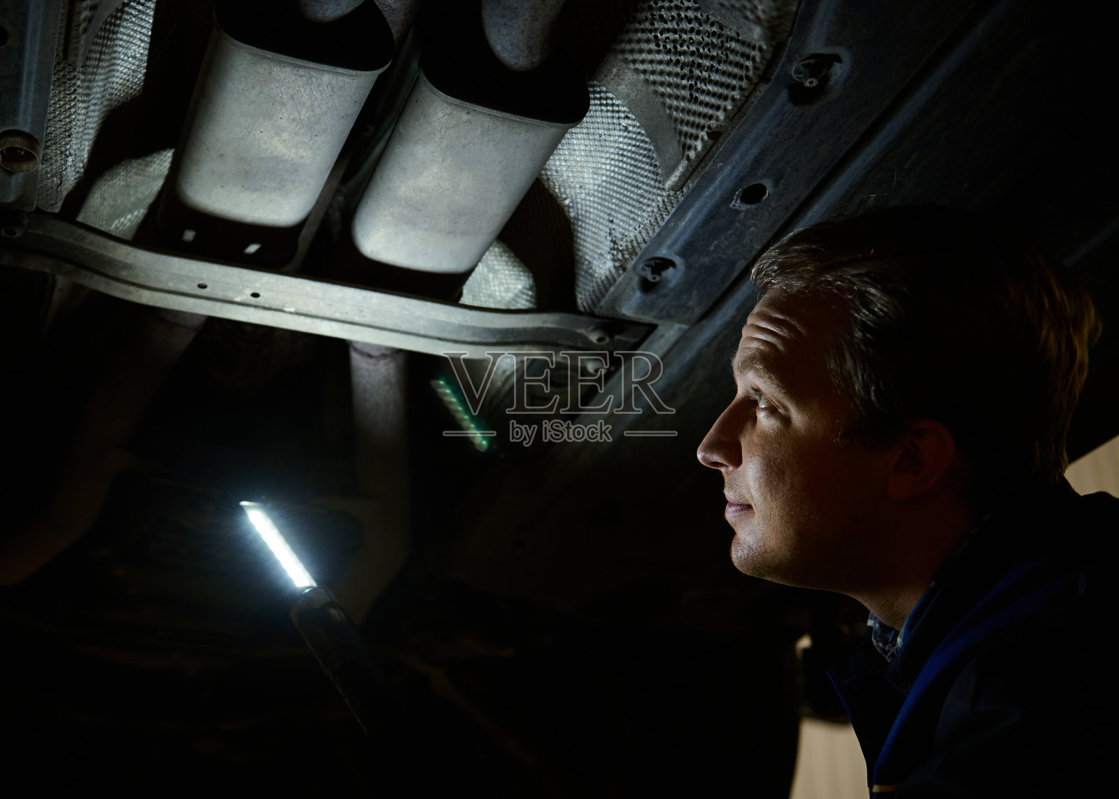 在汽车修理厂检查现代汽车的技术员。在汽车服务中,汽车修理工用手电灯检查起重机上抬起的汽车的底部。汽车维修的概念照片摄影图片