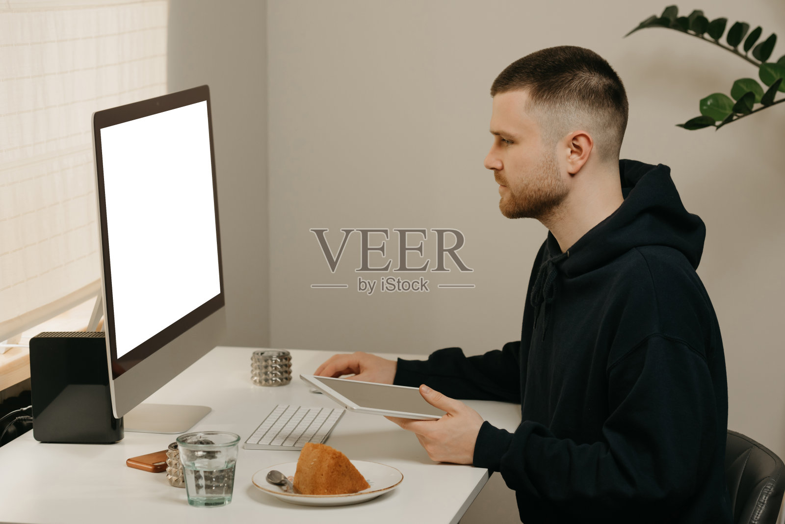 一名男子在家手持一台平板电脑,使用台式电脑远程工作。照片摄影图片