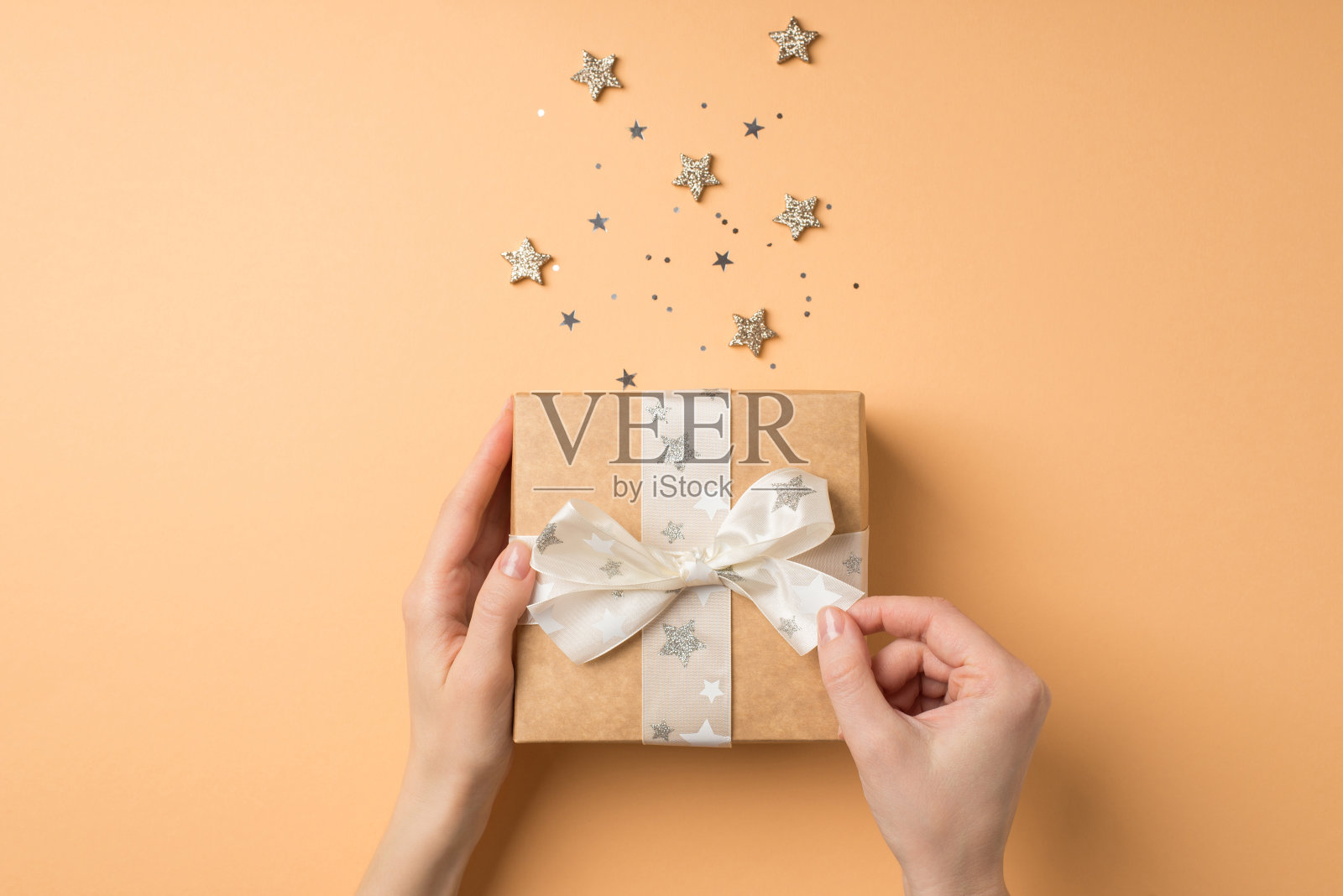 第一人称俯视图照片的情人节decor女性手解开白色蝴蝶结在牛皮纸礼品盒发光的星星和星形纸屑孤立的米色背景照片摄影图片