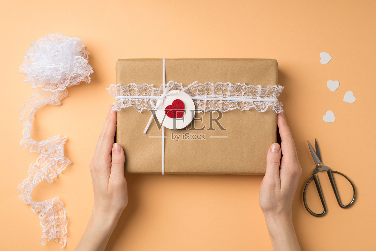 第一人称顶视图照片的情人节装饰女人的手拿牛皮纸礼品盒与花边装饰标签与红色心形剪刀标签线轴的蕾丝在孤立的米色背景照片摄影图片
