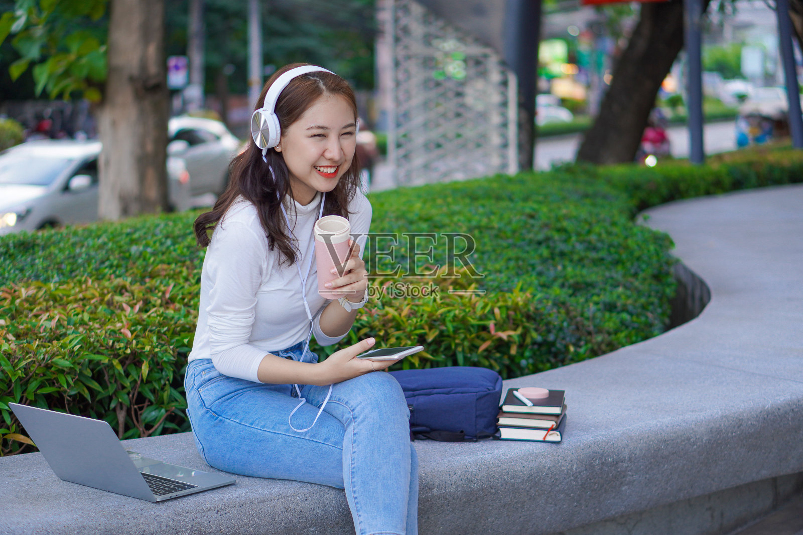 一名女大学生在模糊的地板上开心地笑着,手里拿着智能手机和耳机,旁边放着一个虎皮笔记本电脑包。户外教育交换生概念照片摄影图片